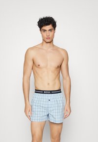 Boxers à carreaux bleu clair avec une taille noire arborant le logo "BOSS", une fermeture à bouton à l'avant et une coupe décontractée.