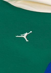 Tecido de malha verde com um acabamento texturizado, apresentando um logo Jumpman bordado em branco no lado esquerdo, acentuado em azul por baixo.