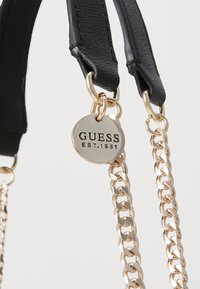 Correa de bolso de imitación cuero negra con acentos de cadena en tono dorado. Presenta una etiqueta metálica redonda grabada con "GUESS EST. 1981."