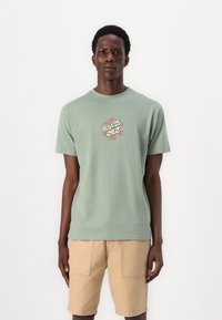 T-shirt de algodão verde claro com um design de logo multicolorido ao centro, decote redondo, mangas curtas e um corte relaxado, combinado com calções bege.