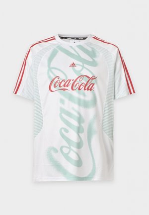 Νέος άνδρας με πλεξούδες, που φοράει λευκό T-shirt Adidas με κόκκινες ρίγες και λογότυπο της Coca-Cola, συνδυασμένο με ανοιχτό μπλε παντελόνια Adidas.