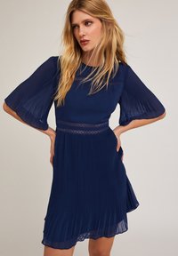 Marineblaues, plissiertes Kleid mit kurzen, ausgestellten Ärmeln und Spitzenakzenten an der Taille. Leichter, strukturierter Stoff mit einem fließenden Design.