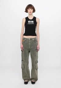 Schwarzes ärmelloses Top mit dem weißen Aufdruck "MOSCHINO JEANS". Olivgrüne Cargo-Hosen mit mehreren Taschen und lockerer Passform. Schwarze Schuhe.