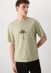 Lys grøn bomuld t-shirt med korte ærmer, der har et centralt grafisk motiv af et palmetræ og tekst nedenunder. Lige kant og afslappet pasform.