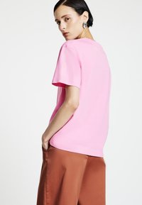T-shirt di cotone rosa con maniche corte e una vestibilità comoda, abbinata a pantaloni marroni a gamba larga. La texture appare morbida e liscia.