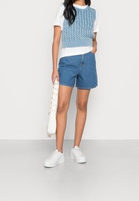 Vaaleansininen neulecropped-paita valkoisen t-paidan päällä, yhdistetty sinisiin farkkushortseihin ja valkoisiin korollisiin tennareihin. Kädessä valkoinen laukku.