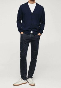 Cardigan bleu marine avec boutons, porté sur un t-shirt blanc, accompagné d'un jean foncé et de baskets blanches avec des accents beige. Design simple et coupe décontractée.