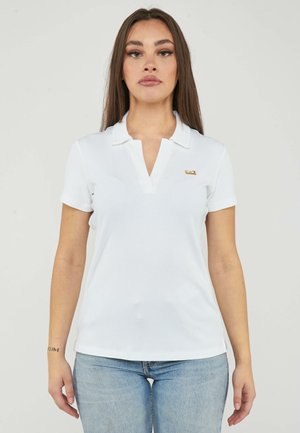 Jonge vrouw met lang bruin haar, die een wit overhemd met korte mouwen en kraag en lichtblauwe spijkerbroek draagt, staat tegen een effen achtergrond.