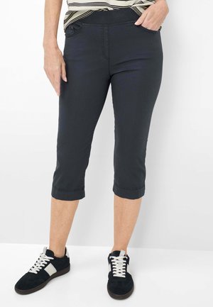 Vrouw in zwarte cropped broek en zwart-witte sneakers, staand met één hand in haar zak.