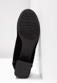 Semelle de chaussure noire avec motif de grip texturé et talon bloc, inclinée contre une surface angulaire blanche.