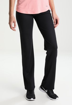 Mujer con pantalones deportivos negros, zapatillas Nike blancas y negras, y una camiseta rosa de Under Armour, de pie contra un fondo blanco.