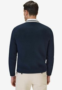 Marinblå stickad tröja med en ribbad textur och en vit randig krage. Har långa ärmar och en figurnära silhuett, bärs över beige byxor.