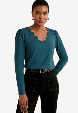Top long à manches longues en teal avec un col festonné et des épaules bouffantes, associé à un jean noir taille haute avec des détails de couture visibles.