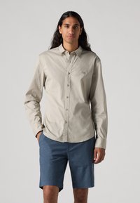 Chemise beige clair à manches longues avec fermeture à boutons et col pointu, associée à un short bleu. Texture lisse, sans motifs ni logos visibles.