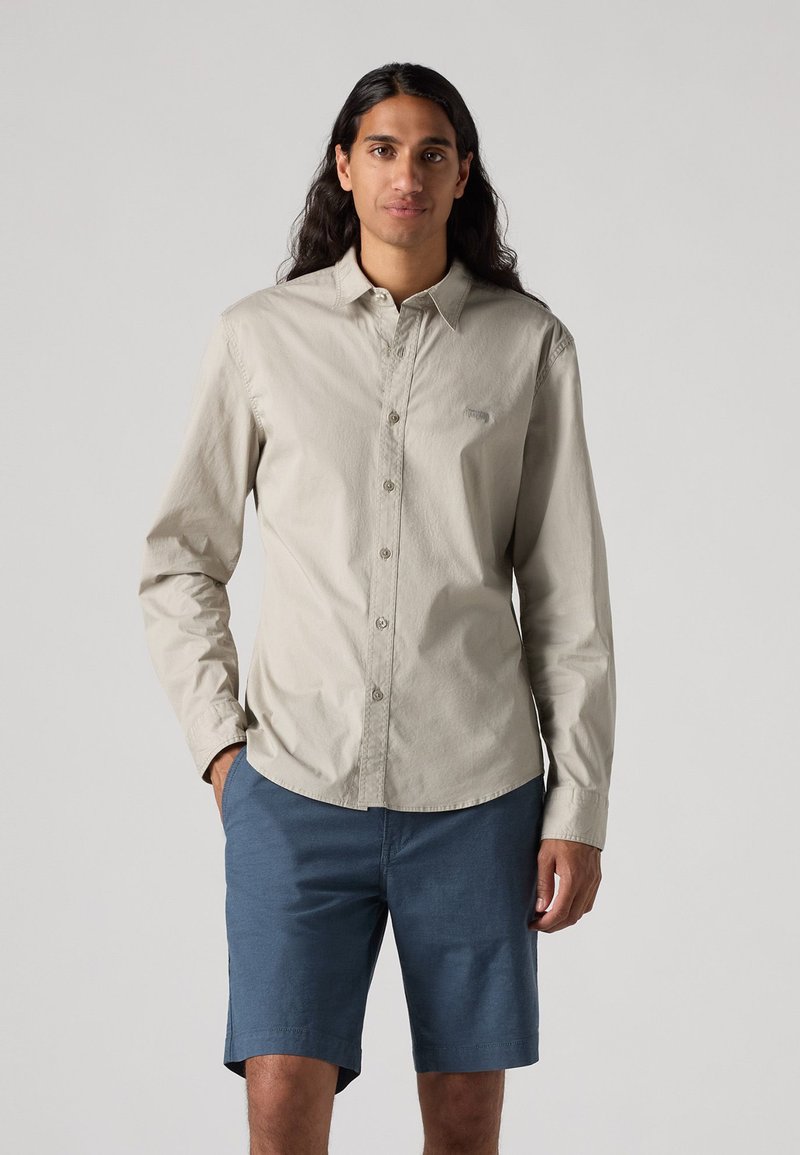 Chemise beige clair à manches longues avec fermeture à boutons et col pointu, associée à un short bleu. Texture lisse, sans motifs ni logos visibles.