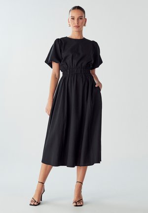 Black dress with short puff sleeves, elastic waistband, and a mid-length flowing skirt. Worn with black strappy heels.