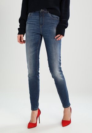 Frau trägt ausgewaschene blaue Skinny-Jeans, kombiniert mit einem schwarzen Pullover und roten High-Heels, steht vor einem schlichten Hintergrund.