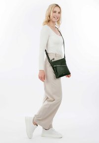 Grüne Leder-Crossbody-Tasche mit strukturierter Oberfläche, ausgestattet mit einer vorderen Reißverschlusstasche und einem verstellbaren Riemen, präsentiert an einem Modell.