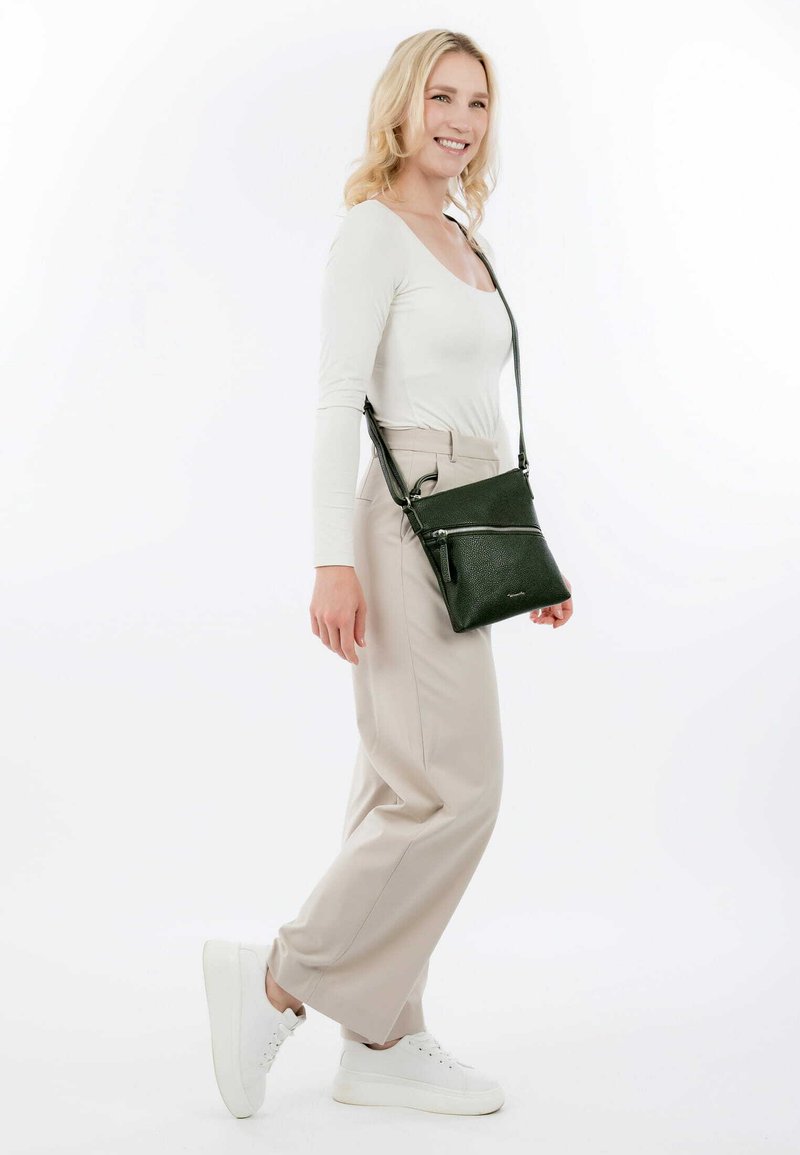 Grüne Leder-Crossbody-Tasche mit strukturierter Oberfläche, ausgestattet mit einer vorderen Reißverschlusstasche und einem verstellbaren Riemen, präsentiert an einem Modell.