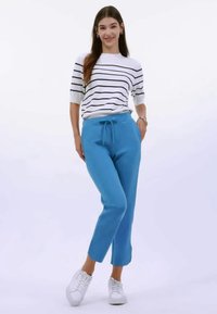 Pantalones jogger azules con cintura ajustable, combinados con un suéter blanco a rayas. El modelo lleva zapatillas blancas y está de pie contra un fondo blanco.