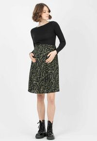 Schwarzes, geripptes Langarmoberteil kombiniert mit einem knielangen, grün gemusterten Rock mit wellenförmigem Design, getragen mit schwarzen Schnürstiefeletten.