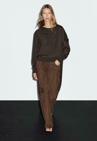 Brown oversized sweater with draped neckline and long sleeves, paired with brown suede wide-leg pants and minimalist black sandals.