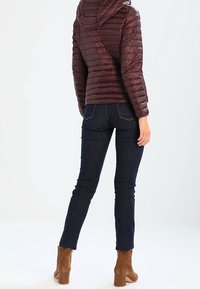 Bordeauxrode donzen jas met capuchon, voorzien van horizontale stiksels en een slank, lichtgewicht ontwerp. Gecombineerd met donkerblauwe spijkerbroek en bruine enkellaarsjes.