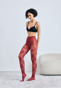 Rote, floral gemusterte Strumpfhosen mit transparenter Textur, kombiniert mit einem schwarzen Bralette, an einem Modell, das neben einem hellen Sofa steht.