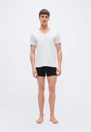 Νέος άνδρας που στέκεται ξυπόλητος, φορώντας λευκό κοντομάνικο μπλουζάκι με V λαιμόκοψη και μαύρο εφαρμοστό σορτς, σε απλό ανοιχτό φόντο.