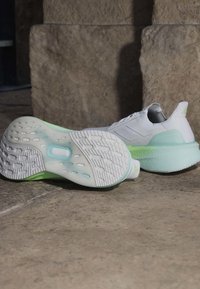 Paire de chaussures de sport blanches et vert clair posées sur un sol en pierre, une chaussure sur le côté montrant la semelle texturée et une autre dressée.