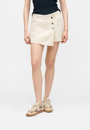 Kobieta ubrana w asymetryczną, kremową mini spódnicę zapinaną na guziki, czarny top bez rękawów oraz beżowe trampki w cętki lamparta.