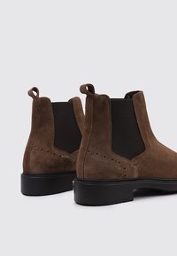 Botas Chelsea de ante marrón con paneles laterales elásticos negros, puntera redonda, sutiles detalles perforados y suelas de goma negra para mayor agarre.