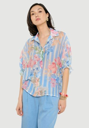 Frau trägt eine transparente, geknöpfte Bluse mit blau-weißen vertikalen Streifen und rosa Blumenmuster, kombiniert mit hellblauen Hosen und Armbändern.