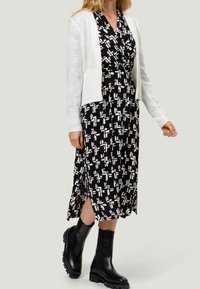 Robe noire avec motif géométrique blanc, longueur genoux, associée à un cardigan court en tricot blanc et des bottes noires à semelle épaisse.