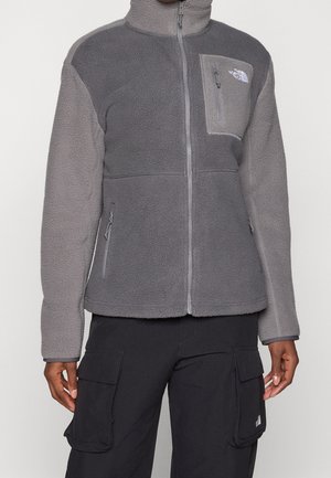 Personne portant une veste polaire grise avec des poches zippées et une poche poitrine, associée à un pantalon cargo noir avec de grandes poches latérales.