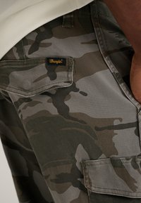 Camo cargo pants in green, gray, and beige fabric, featuring a Wrangler logo tag and a side pocket with flap closure. Smooth texture.
