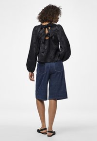 Blusa nera con texture e chiusura con cravatta sul retro, abbinata a pantaloni shorts di denim a lunghezza ginocchio. Il modello è in posizione laterale, mostrando sandali semplici.