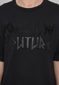 Melna T-kreklā ar reljefu izceltu "VERSACE JEANS COUTURE" logo dažādās tekstūrās un formās. Apaļš kakls, īsas piedurknes.