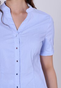 Chemise bleu clair à manches courtes, à rayures blanches verticales et col mandarin. Dispose de petits boutons métalliques sur le devant.