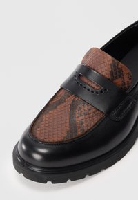 Sorte skinnloafers med en brun slange-mønstrete tekstiloverdel, med en dekorativ stropp med perforeringer og en robust såle.
