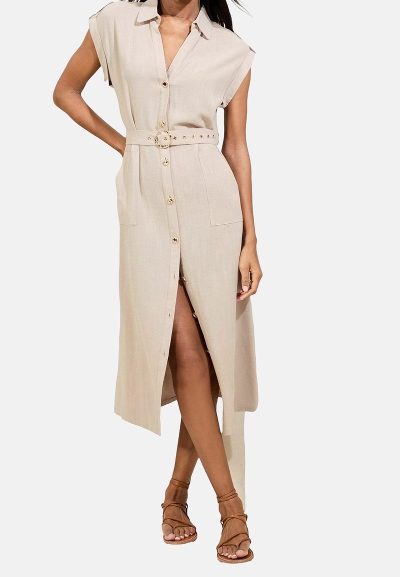Femme portant une robe midi beige sans manches boutonnée avec une ceinture à la taille et des sandales marron à brides, posant avec une main sur la hanche.