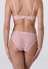 Donna che indossa un reggiseno di pizzo rosa e slip coordinati in pizzo, vista da dietro, con piccoli tatuaggi sul braccio e sulla parte superiore della schiena.
