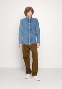 Camicia di jeans azzurro chiaro con colletto button-down, taschino frontale e maniche lunghe, abbinata a pantaloni marroni e sneaker bianche.