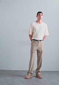 Beżowy, przewiewny t-shirt z prążkowanej tkaniny, khaki spodnie cargo z bocznymi kieszeniami, w parze z jasnobrązowym obuwiem. Neutralne tło.