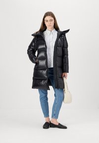 Zwarte pufferjas met een rits aan de voorkant, capuchon en zijzakken; heeft een glanzende, gewatteerde textuur en een lange snit; gestyled over een gestreept shirt.
