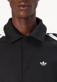 Casaco preto com botão na frente e colarinho, apresentando realces brancos nos ombros e um pequeno logotipo bordado em branco no peito. Textura suave.