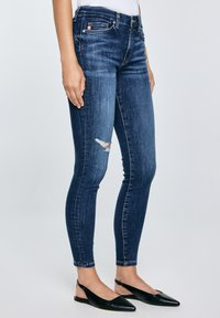 Plonas tamsiai mėlynas džinsas su mažyte plyšiu šlaunyje, dėvimas stovinčio žmogaus, derinamas su juodais aštriapadžiais slingback batais.