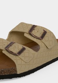 Sandale beige avec des brides en tissu texturé, deux boucles en bronze gravées de "ROXY,", semelle intérieure en liège et semelle noire visible sur fond blanc.