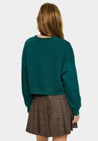 Teal cropped sweatshirt met verlaagde schouders en geribbelde manchetten, gecombineerd met een bruine geruite rok met kleurrijke spikkels.