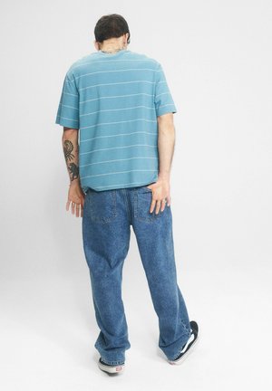 Mężczyzna z tatuażami, ubrany w niebieski, w paski t-shirt, luźne niebieskie jeansy i czarne trampki, stoi tyłem na białym tle.